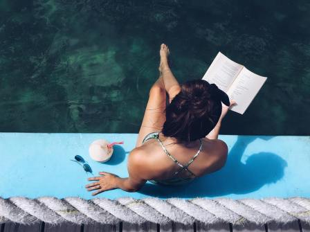 Young woman sits on the side of a pool with a book in hand