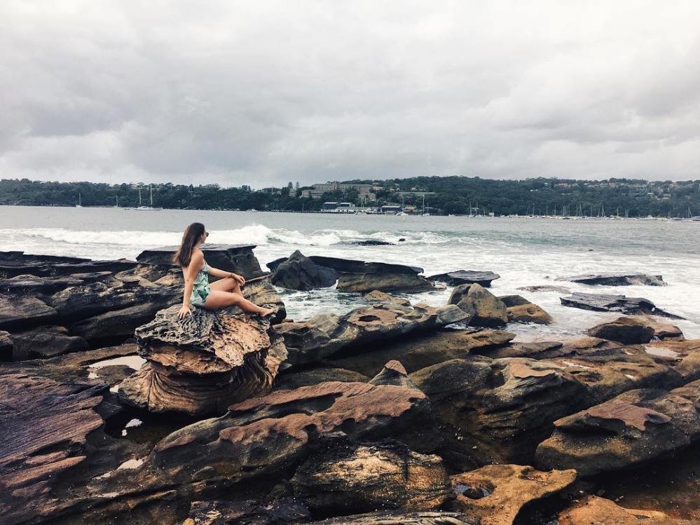 Woman wearing a floral swimsuit sitting on the rocky foreshore of Balmoral Beach in Sydney, Australia