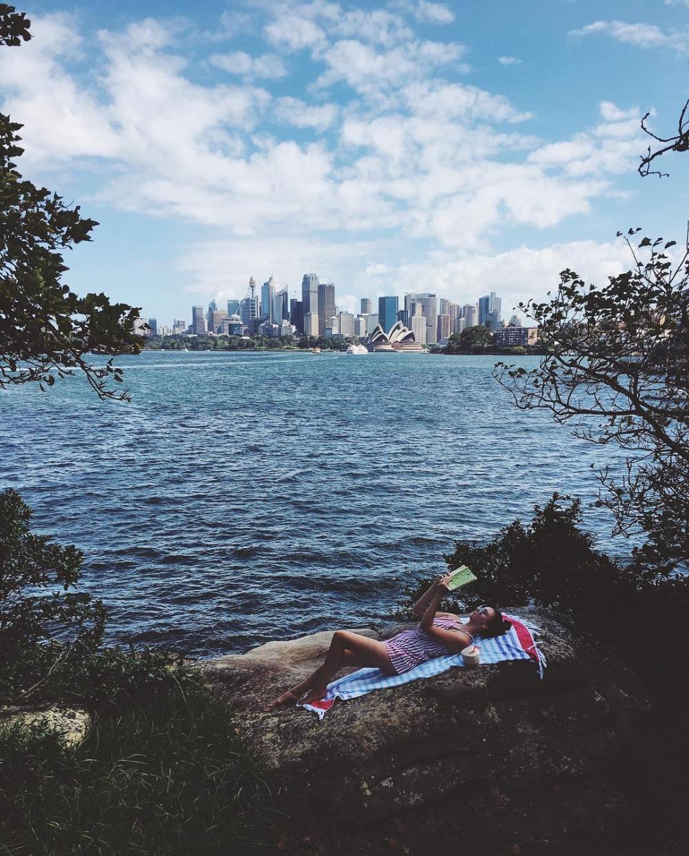 Woman lays on a rock with a book in her hands, the Sydney skyline in the background