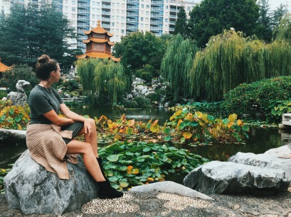 Woman sits on a rock with a pond and temple in the background in the Chinese Garden of Friendship