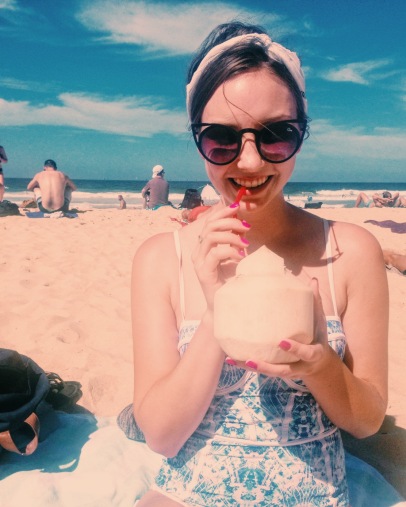 Young woman wearing sunglasses and a bandana sips from a coconut on Manly Beach