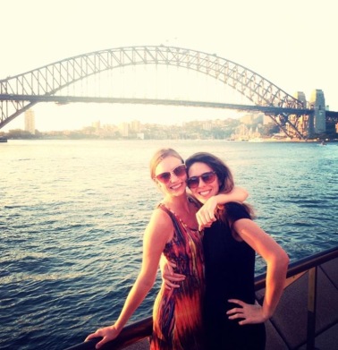 Two young woman smile at the camera in front of the Harbour Bridge